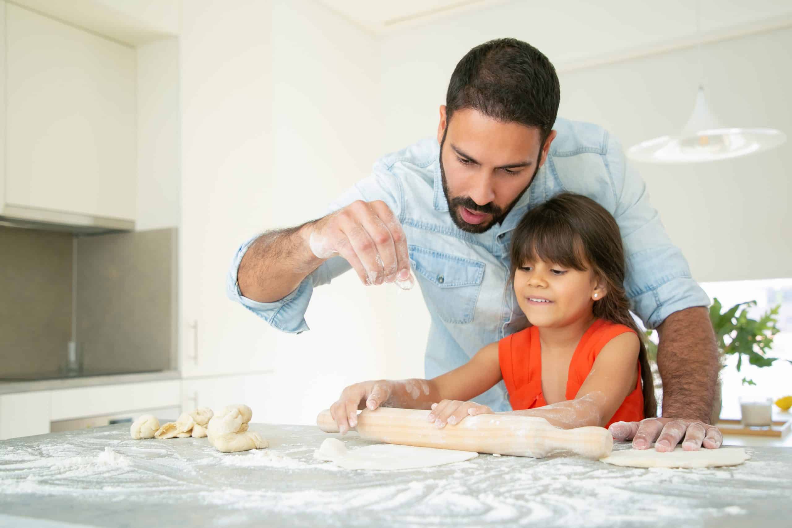 Cheerful girl and her dad kneading and rolling dough